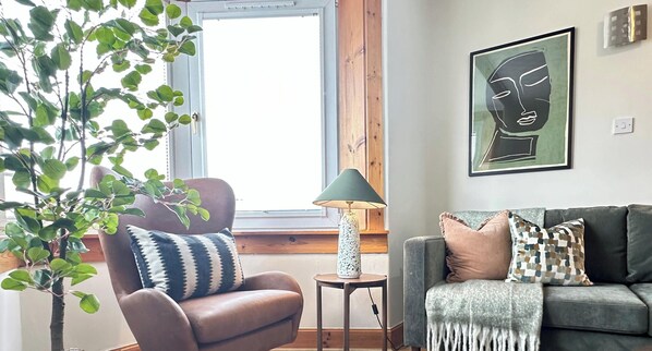 A living room with a brown leather armchair, a grey sofa, a green floor lamp, and a wall-mounted abstract art piece.