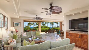 A spacious living area with a large sofa, a coffee table, and a television. There are ceiling fans, a dining area with chairs, and a view of palm trees and the ocean. - Elevated 2BR Hillside Villa 4102 at Hualalai – Ocean Views & Island Lifestyle (Kailua Kona)