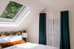 Habitación de hotel con cama, mesita de noche, lámpara, ventana con cortinas y vista a zonas verdes.