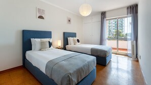 Une chambre d’hôtel avec deux lits, une grande fenêtre donnant sur des immeubles, un luminaire au plafond et un climatiseur fixé au mur.