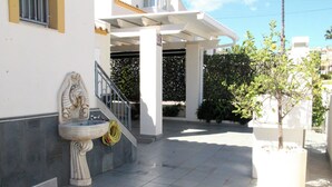 Un edificio con fachada blanca, una fuente de piedra, un árbol en maceta y una escalera cubierta.