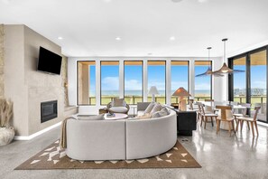 A modern living room with a large sectional sofa, a flat-screen TV mounted on the wall, and a dining area with a table and chairs. - Sandbox: Modern Home with Pool, Spa, Elevator, Direct Beach Access (Oak Island)