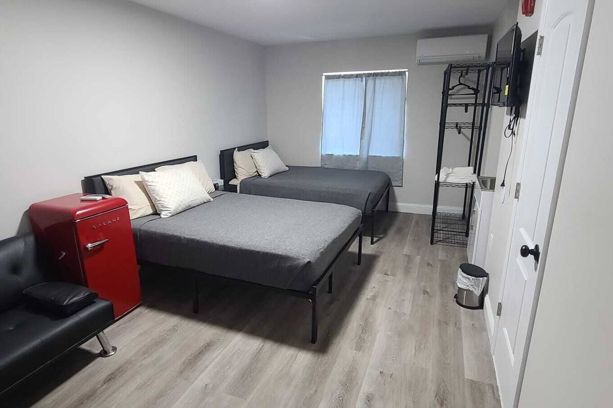 Une chambre de taille modeste, équipée de deux lits, d’un mini-bar rouge, d’un canapé noir et d’une fenêtre avec des rideaux.