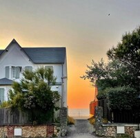 Una casa costera con un sendero de piedra que lleva al mar, rodeada de vegetación y una valla de madera.