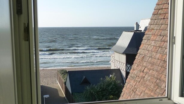 A view from a window overlooking a beach with waves, a building with a sloped roof, and a clear sky. - Hotel de la Mer (Blonville-sur-Mer)