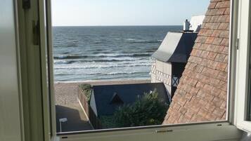 Blick aus einem Fenster auf einen Strand mit Wellen, ein Gebäude mit schrägem Dach und einem klaren Himmel.