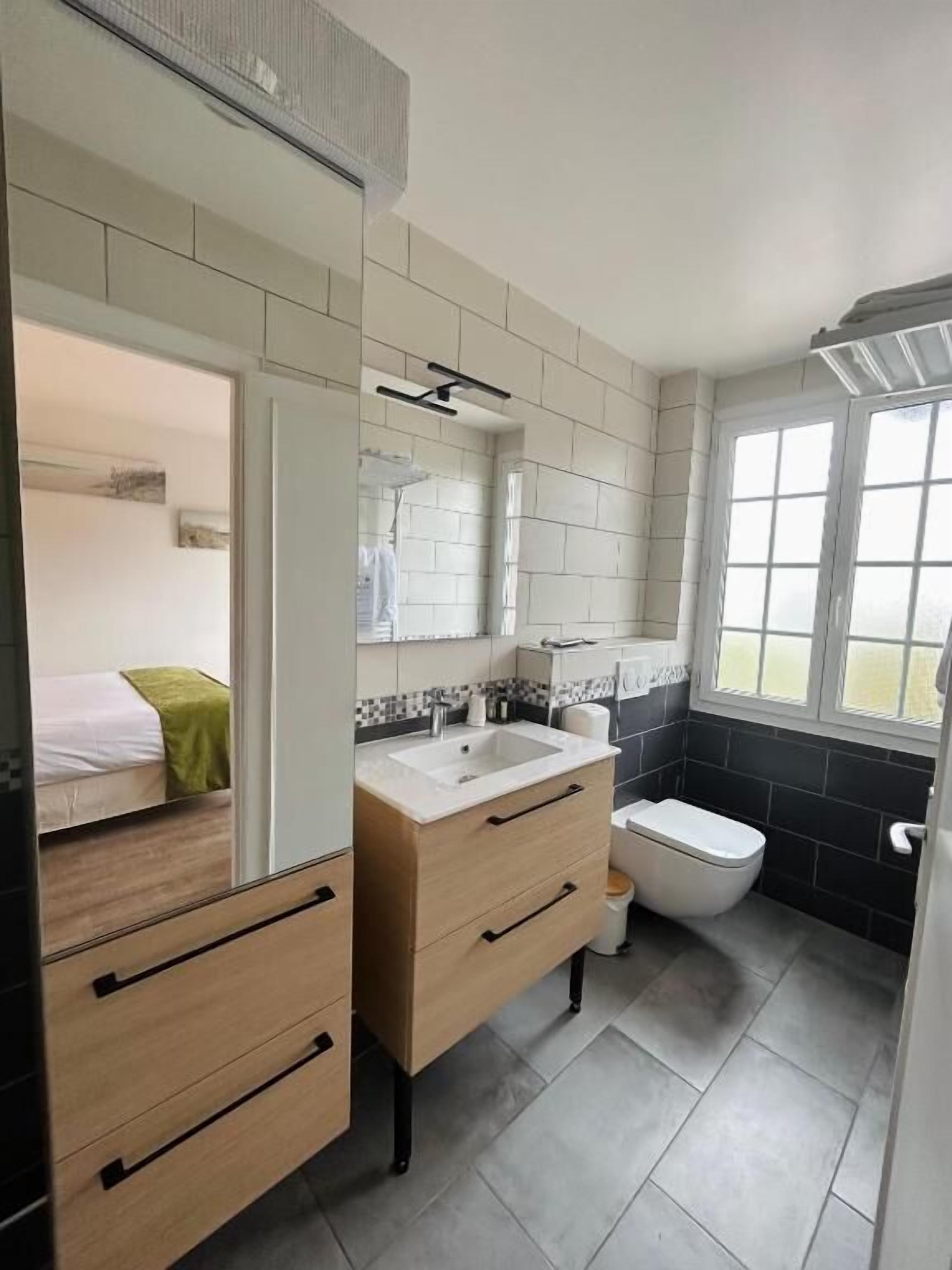 A modern bathroom with a double sink, a toilet, and a shower area. There is a bed visible in the adjacent room.