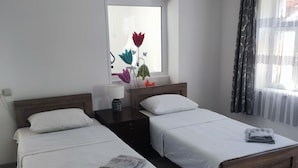 Une chambre d’hôtel avec deux lits en bois, du linge de lit blanc, une table de chevet avec une lampe et une fenêtre ornée de fleurs décoratives.