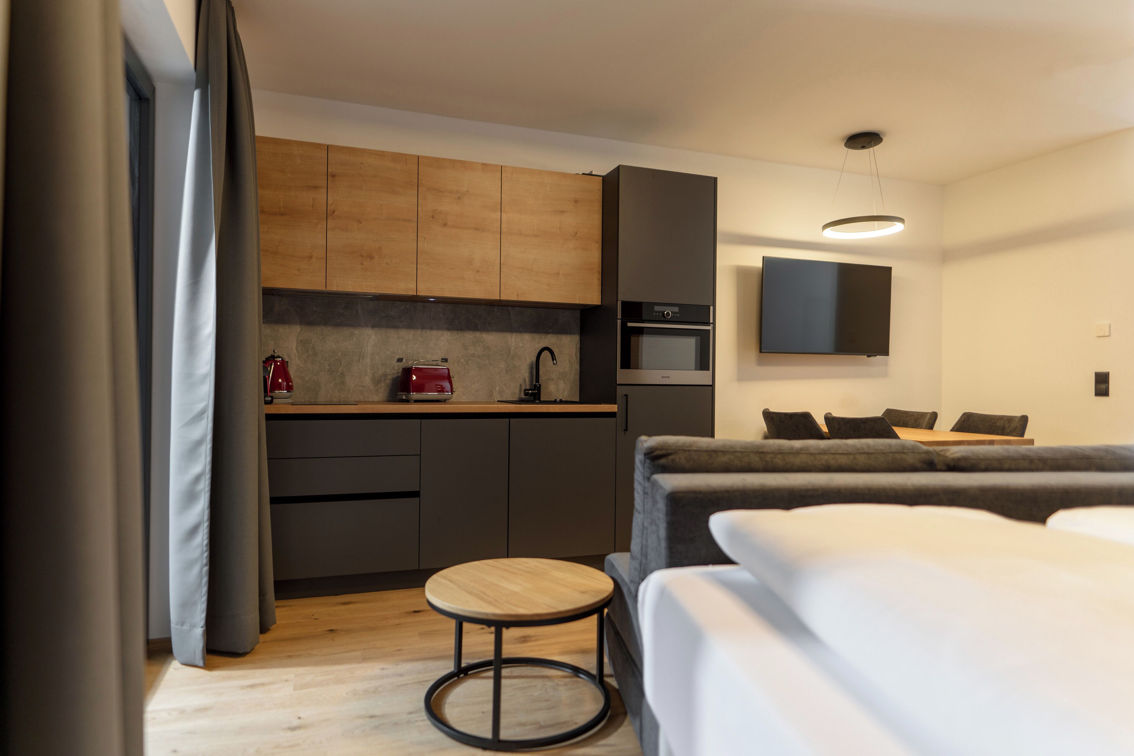 A compact hotel room with a kitchenette, a television, a sofa, and a small round table.