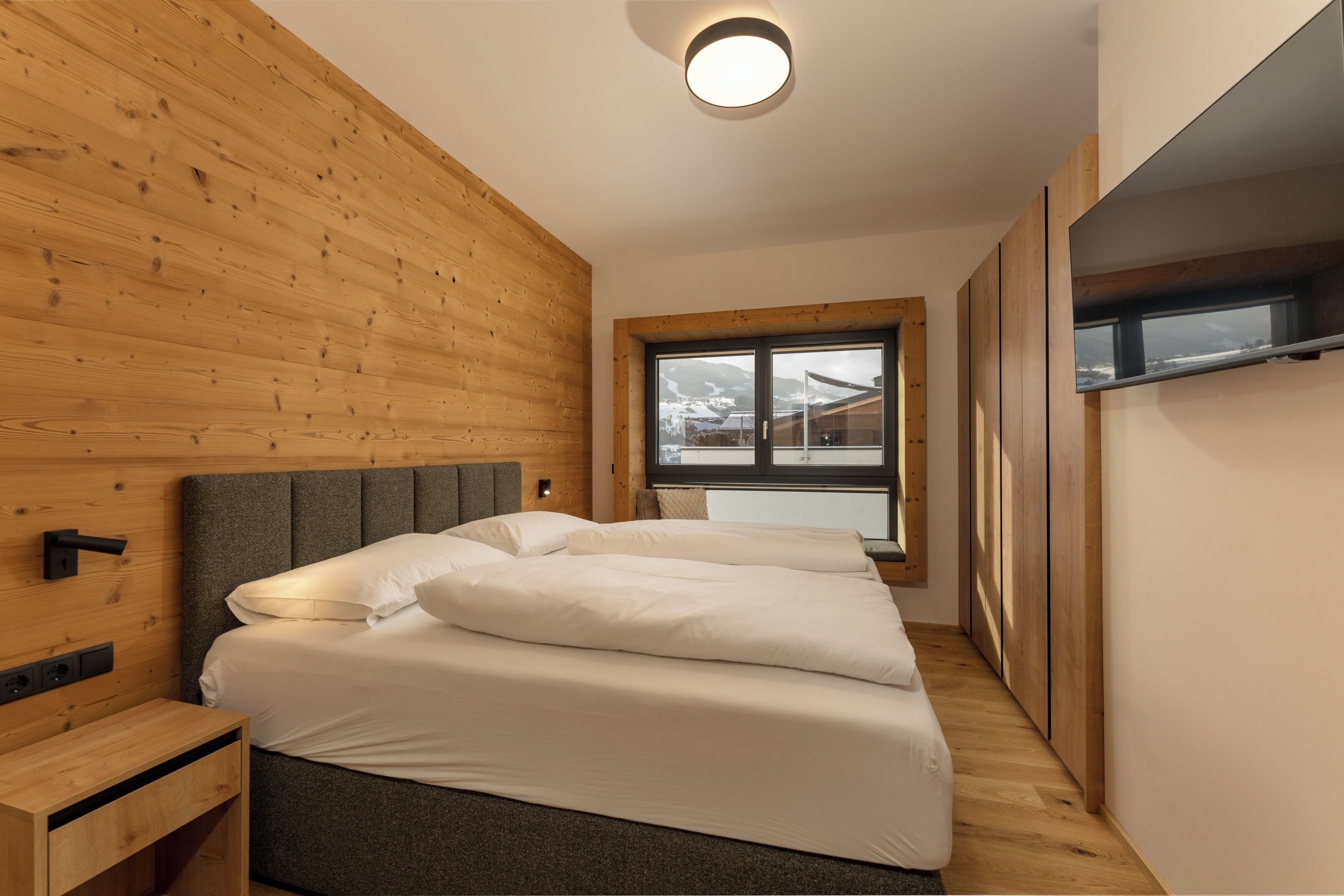 Un dormitorio con cabecera de madera, cama con sábanas blancas, una mesita de noche y una ventana con vistas a un paisaje nevado.