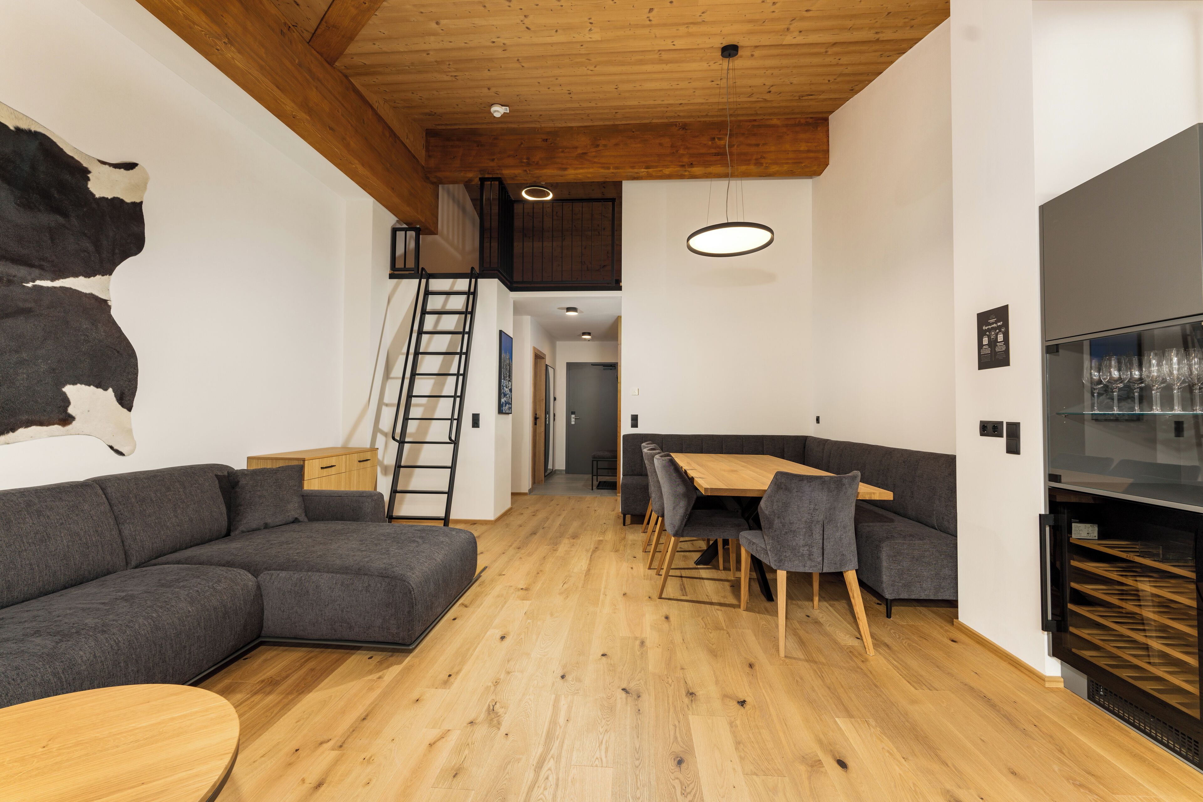 Een moderne woonkamer met een houten plafond, een grijze modulaire bank, een houten eettafel met stoelen, en een zwarte koelkast.