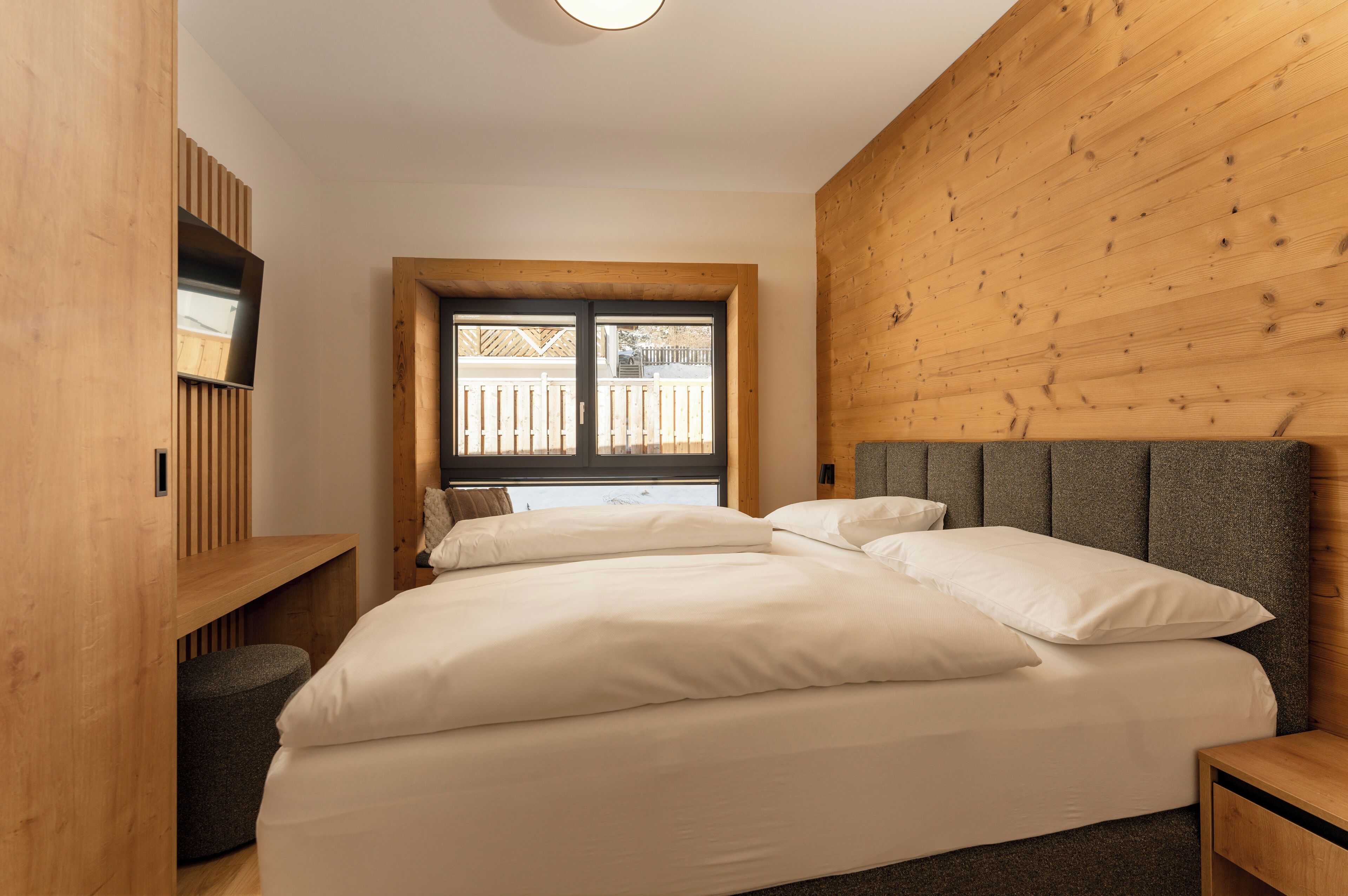 A bedroom with a bed, a wooden headboard, a TV mounted on a wooden panel, and a window with a view of a wooden deck.