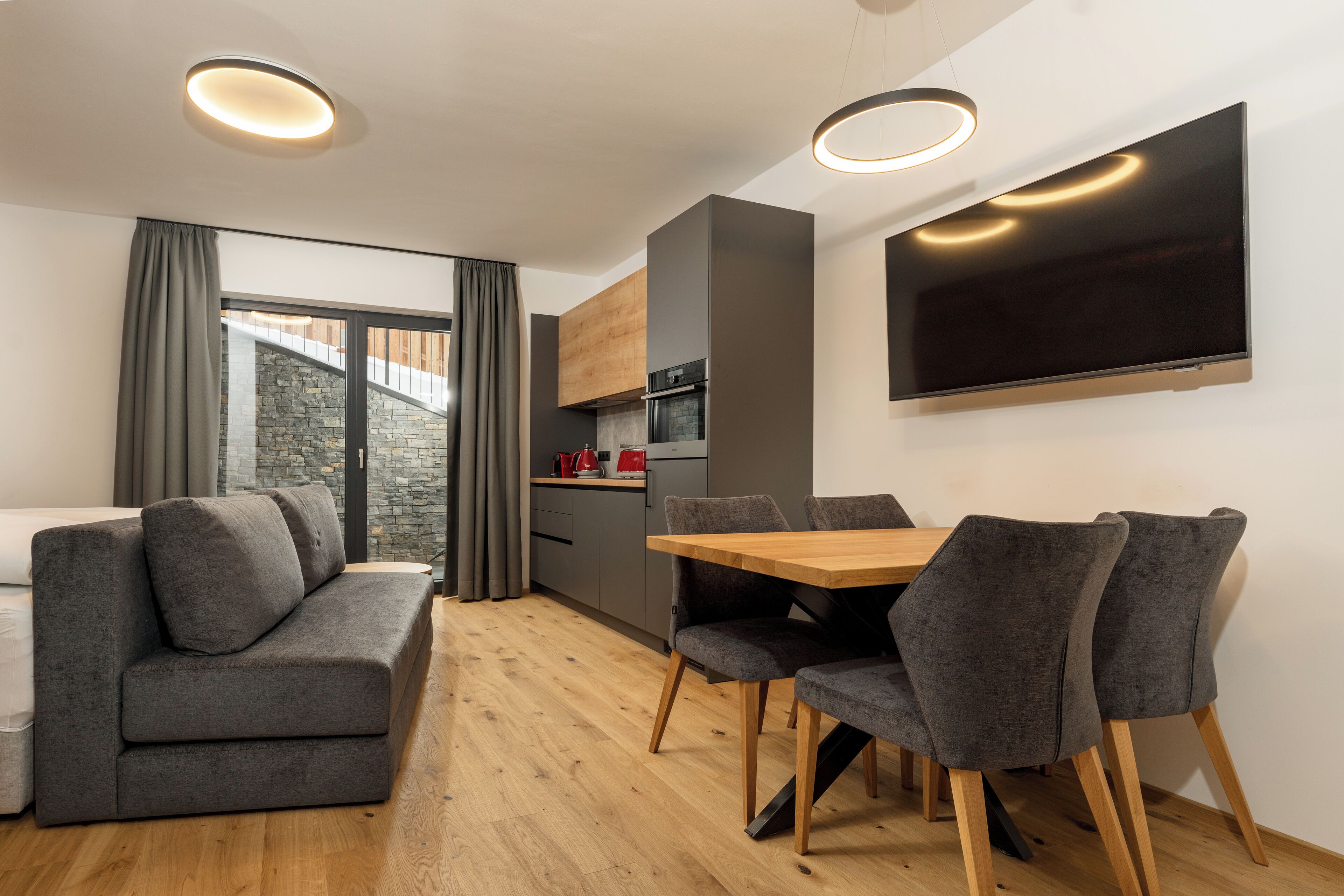 A modern living room with a sofa, dining table, and chairs, a kitchen area, and a large TV mounted on the wall.