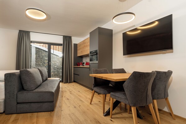 A modern living room with a sofa, dining table, and chairs, a kitchen area, and a large TV mounted on the wall. - Pure Alpine Suites (Schladming)