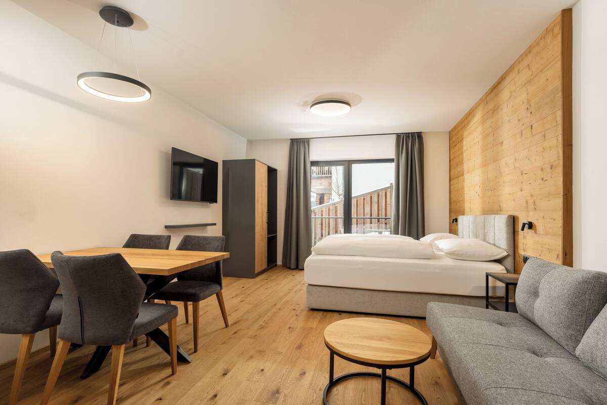 Une chambre d’hôtel moderne avec un plancher en bois, un lit, un canapé, une table à manger avec des chaises, une télévision et un balcon avec vue.