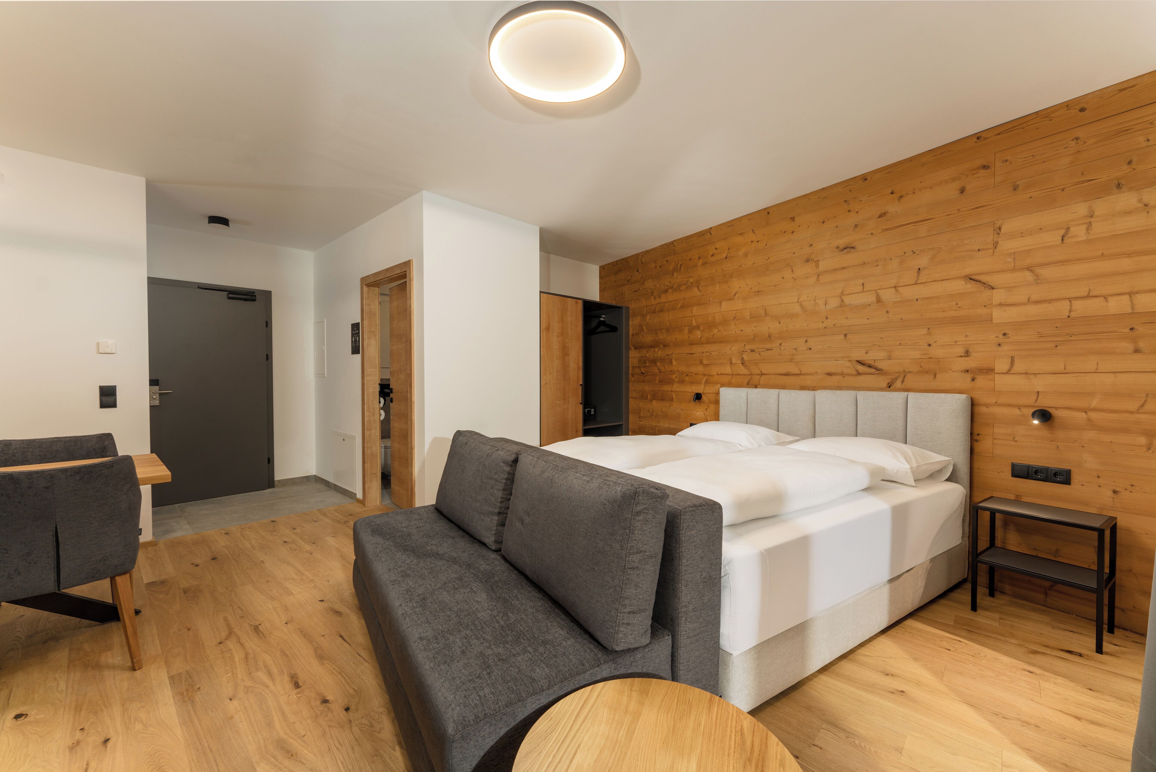 Un dormitorio moderno con una cama grande, un sofá gris, una mesita pequeña y paneles de madera en las paredes.