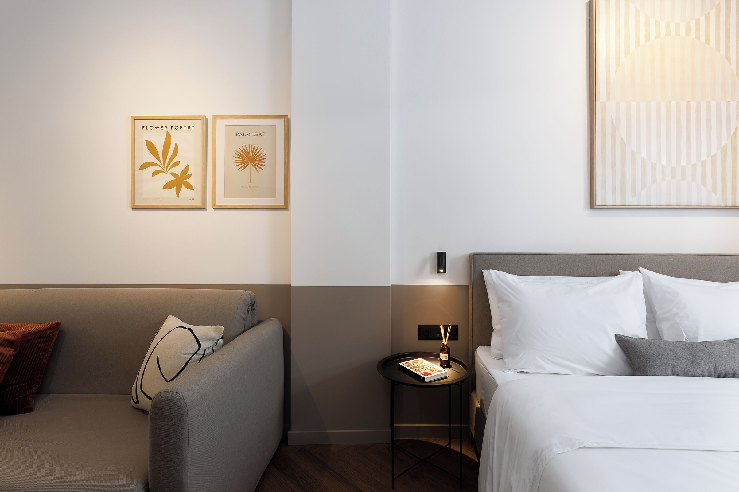 A hotel room with a bed, a sofa, a small table with a book, and two framed artworks on the wall.