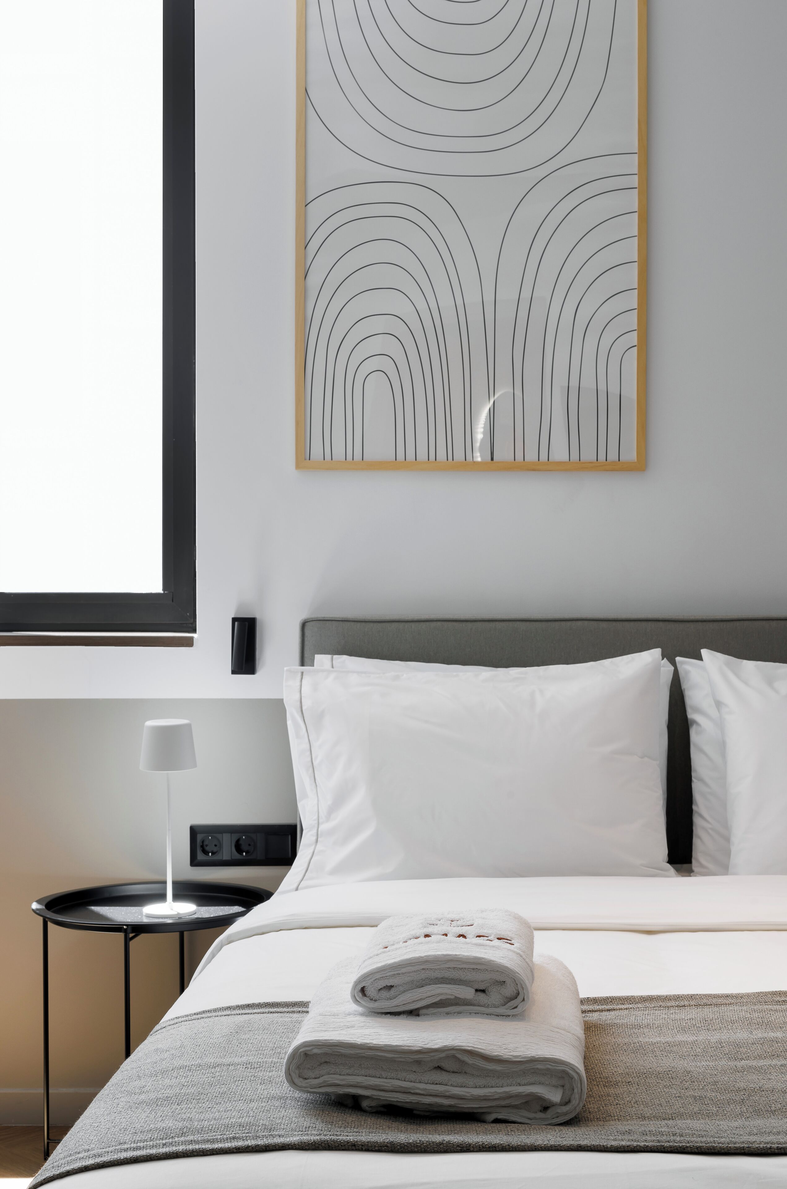 A neatly made bed with white linens, a grey blanket, and a folded towel on a grey bedspread. A black nightstand with a white lamp is beside the bed.