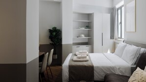 A modern hotel room with a neatly made bed, a seating area with a chair and a table, a potted plant, and built-in shelving. - ERMASS Athens (Athens)