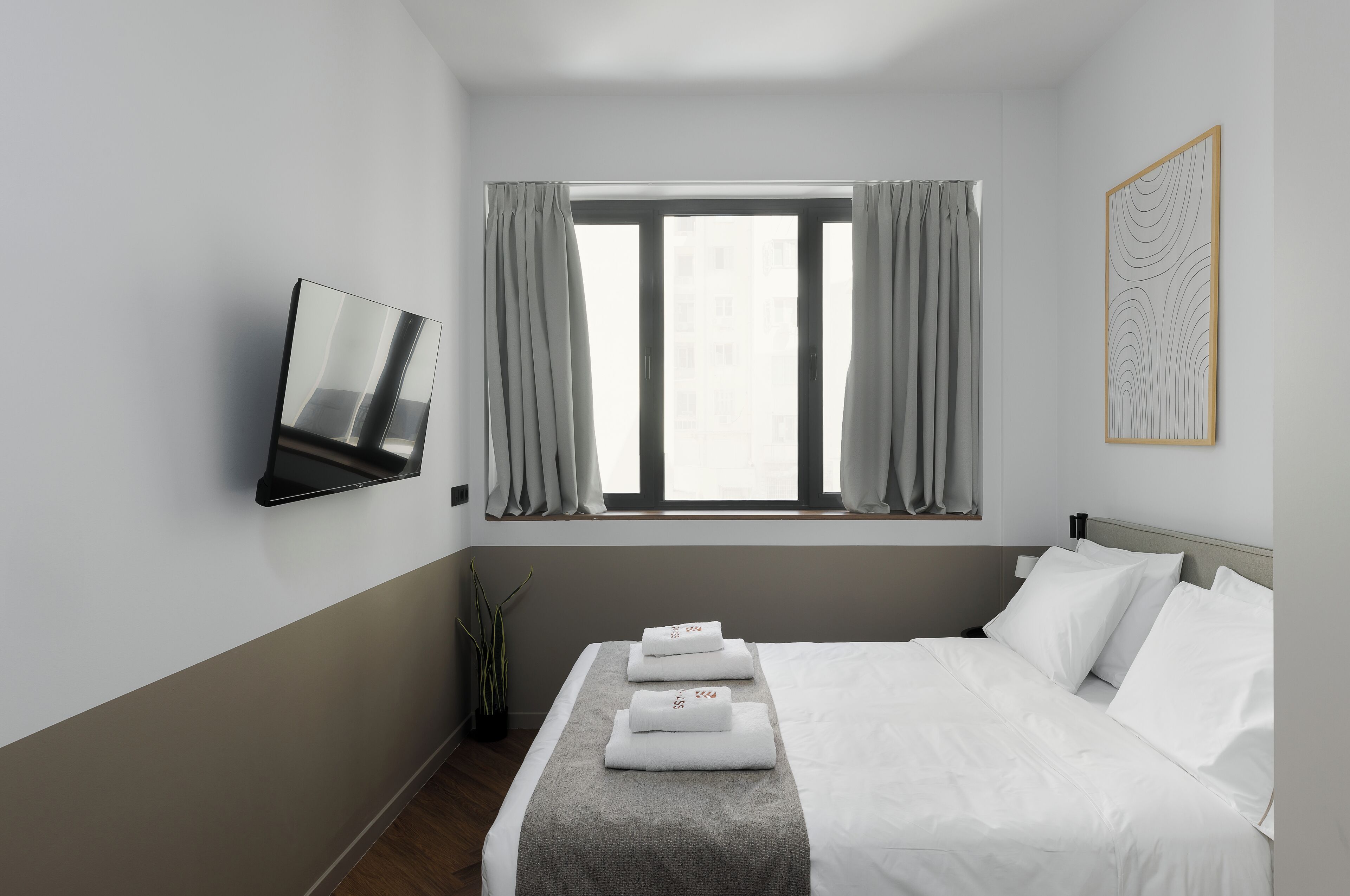 Ett hotellrum med en säng, en tv som är monterad på väggen, ett fönster med gardiner och ett inramat konstverk på väggen.