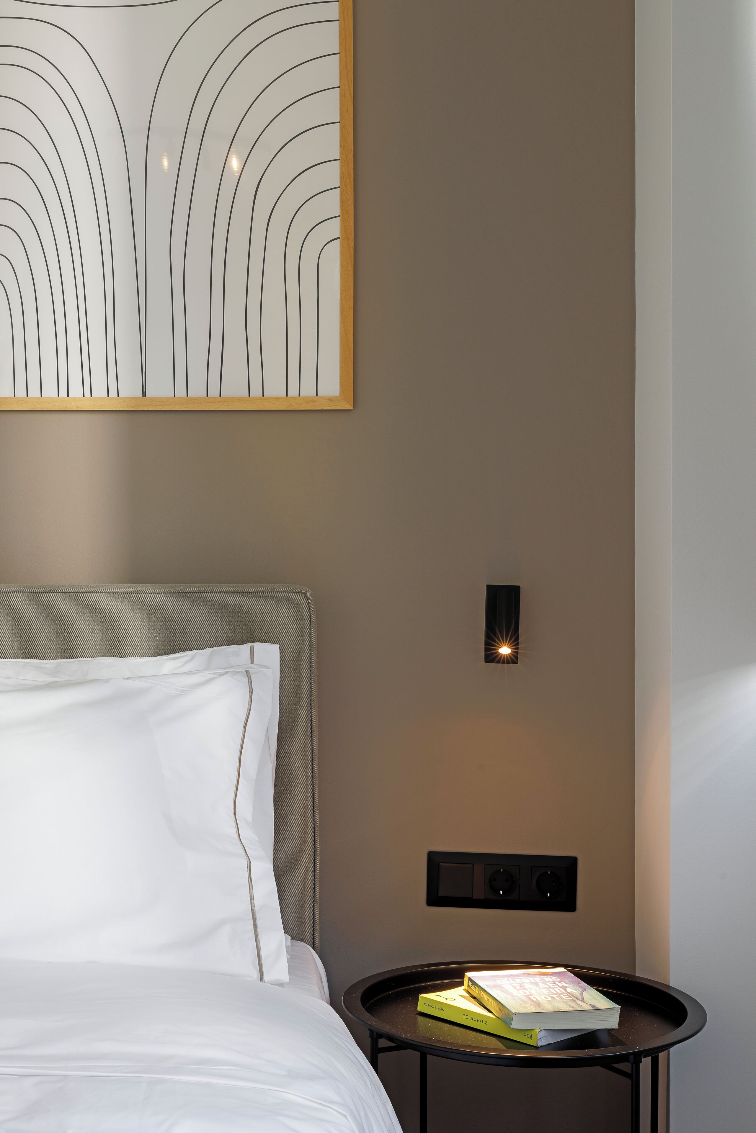 Una cama bien tendida con sábanas blancas, una mesita redonda con un libro y una obra de arte moderna sobre la cama.
