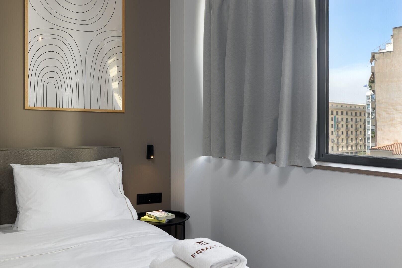 A hotel room with a bed, a nightstand, a wall art piece, a window with curtains, and a view of buildings outside.