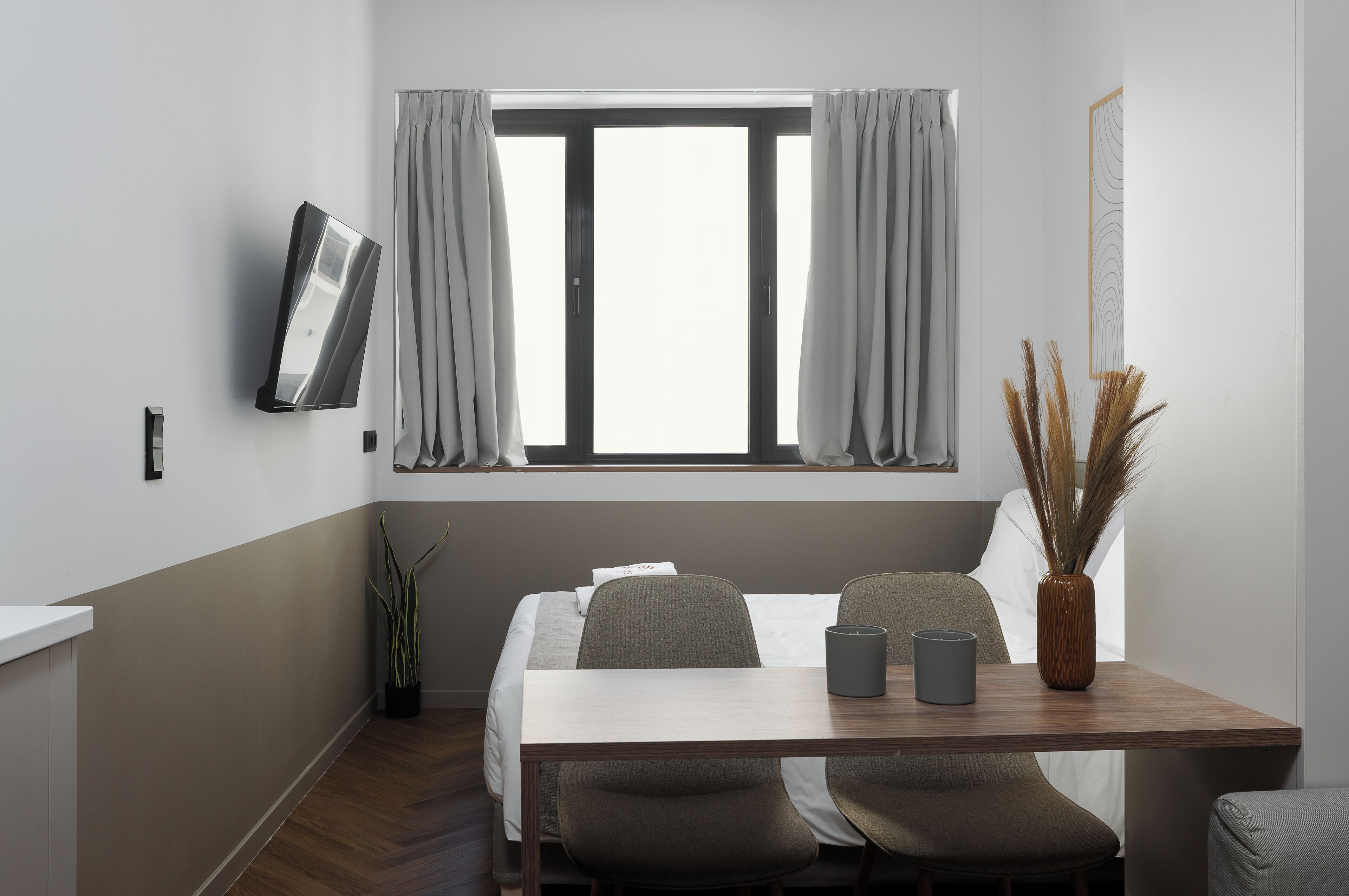 A modern hotel room with a bed, a desk with two chairs, a vase with dried plants, and a window with curtains.