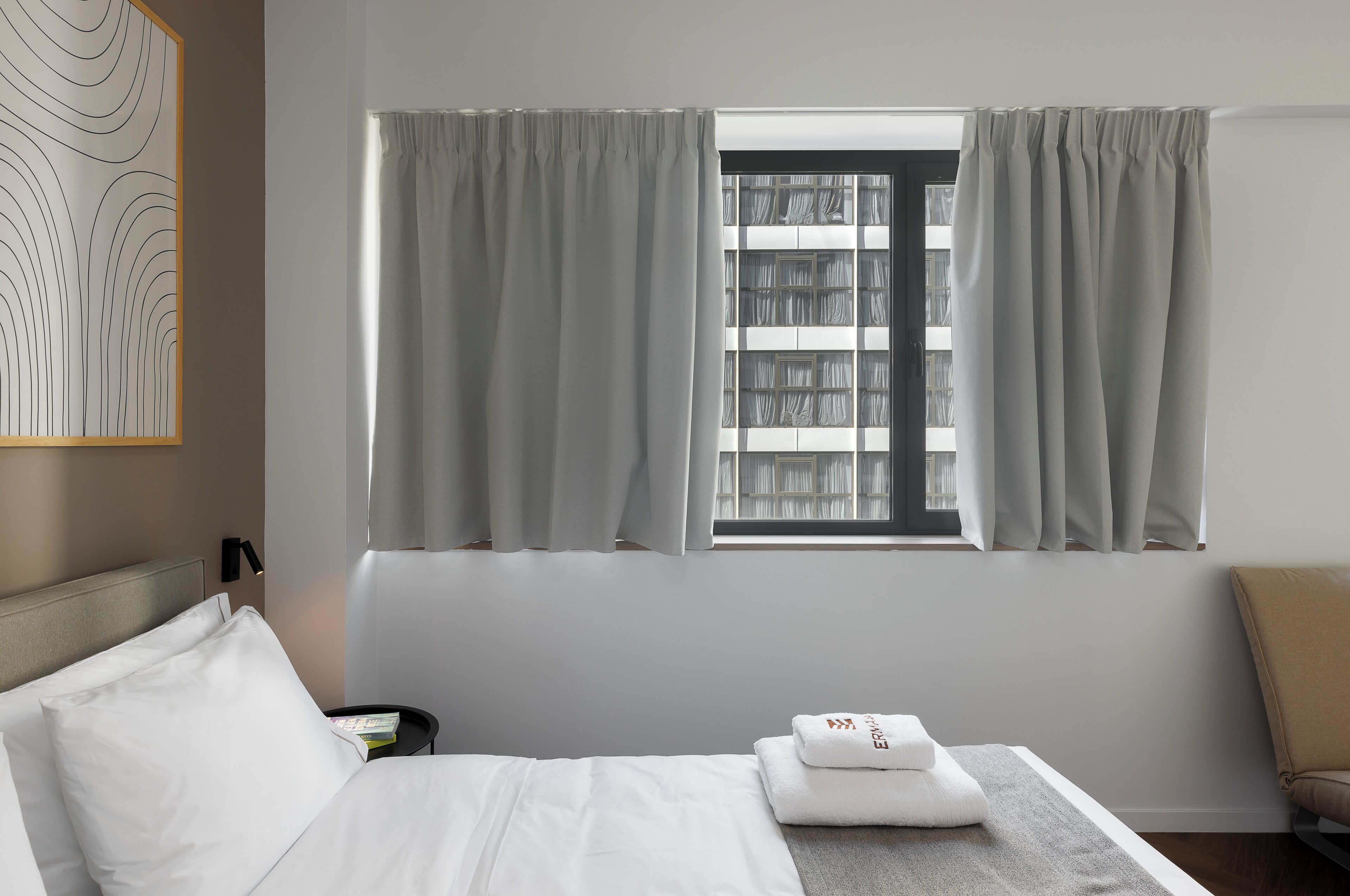 Hotellihuone, jossa on sänky, yöpöytä, verhoilla varustettu ikkuna, sängynpääty ja seinälle kiinnitetty taideteos.