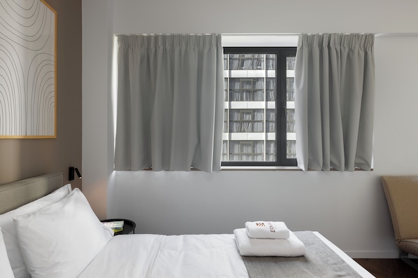 Una habitación de hotel con cama, mesita de noche, ventana con cortinas, cabecera y una obra de arte colgada en la pared.