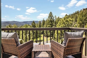 A balcony with wicker furniture and a table, overlooking a forested area with mountains in the distance.