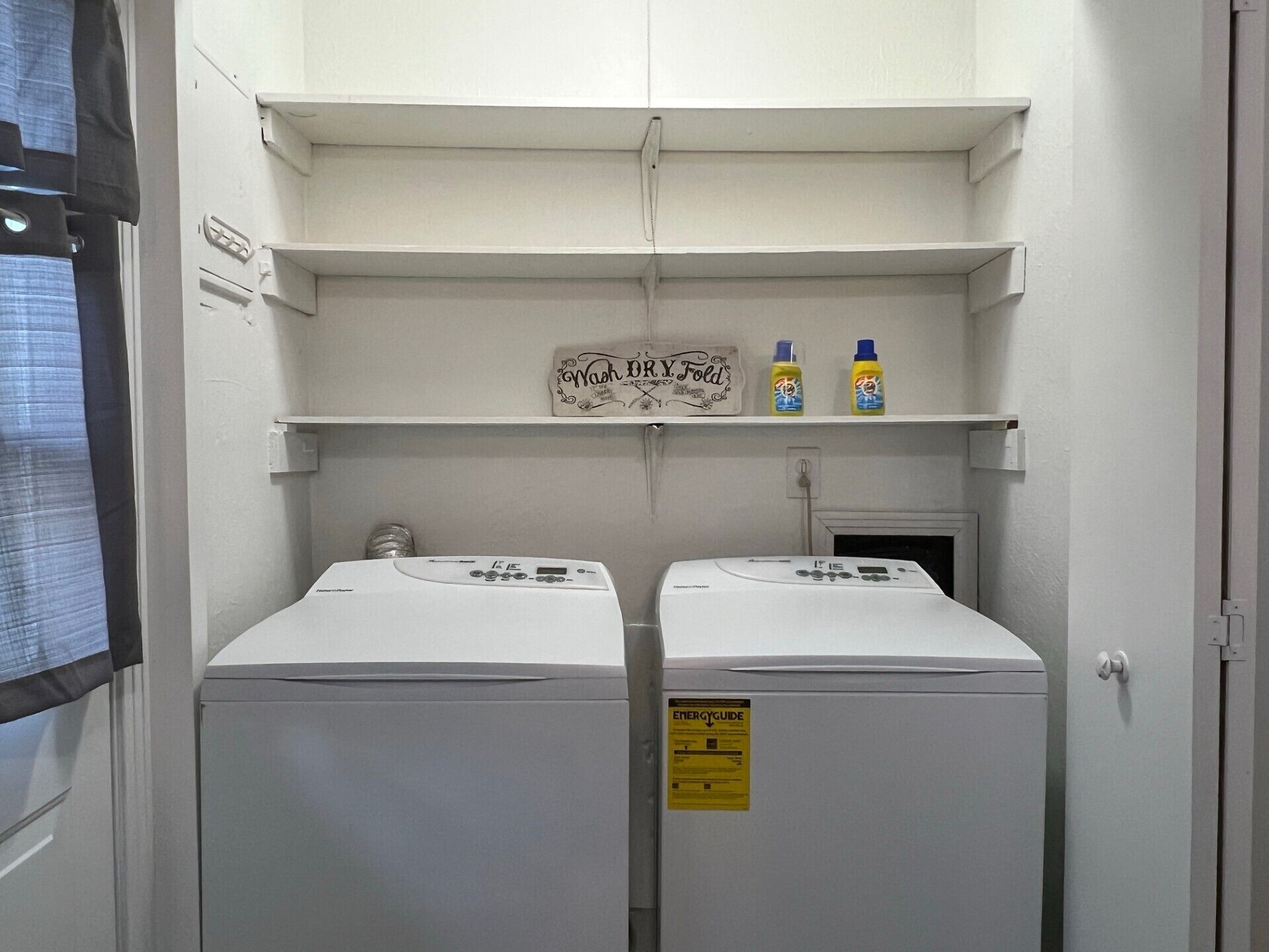 A laundry room with two washing machines and a shelf above containing two bottles of detergent.