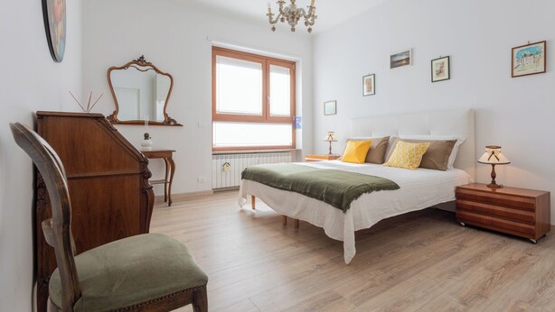 Un dormitorio con cama, mesita de noche, lámpara, silla, cómoda, ventana y candelabro.