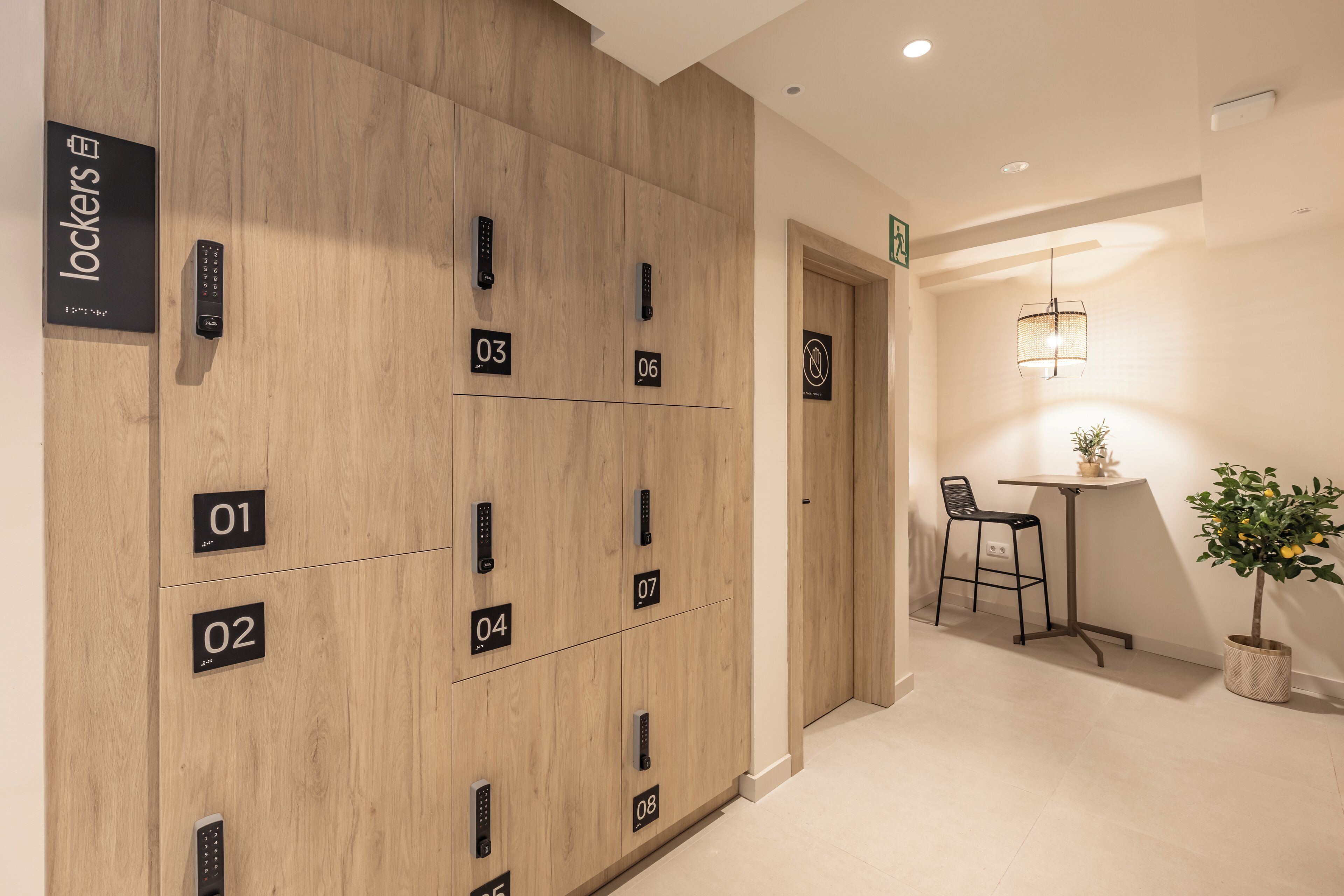 A hotel room with a wooden panel wall, numbered lockers, a small table with a chair, and a potted plant.