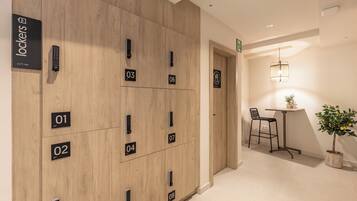 A hotel room with a wooden panel wall, numbered lockers, a small table with a chair, and a potted plant.