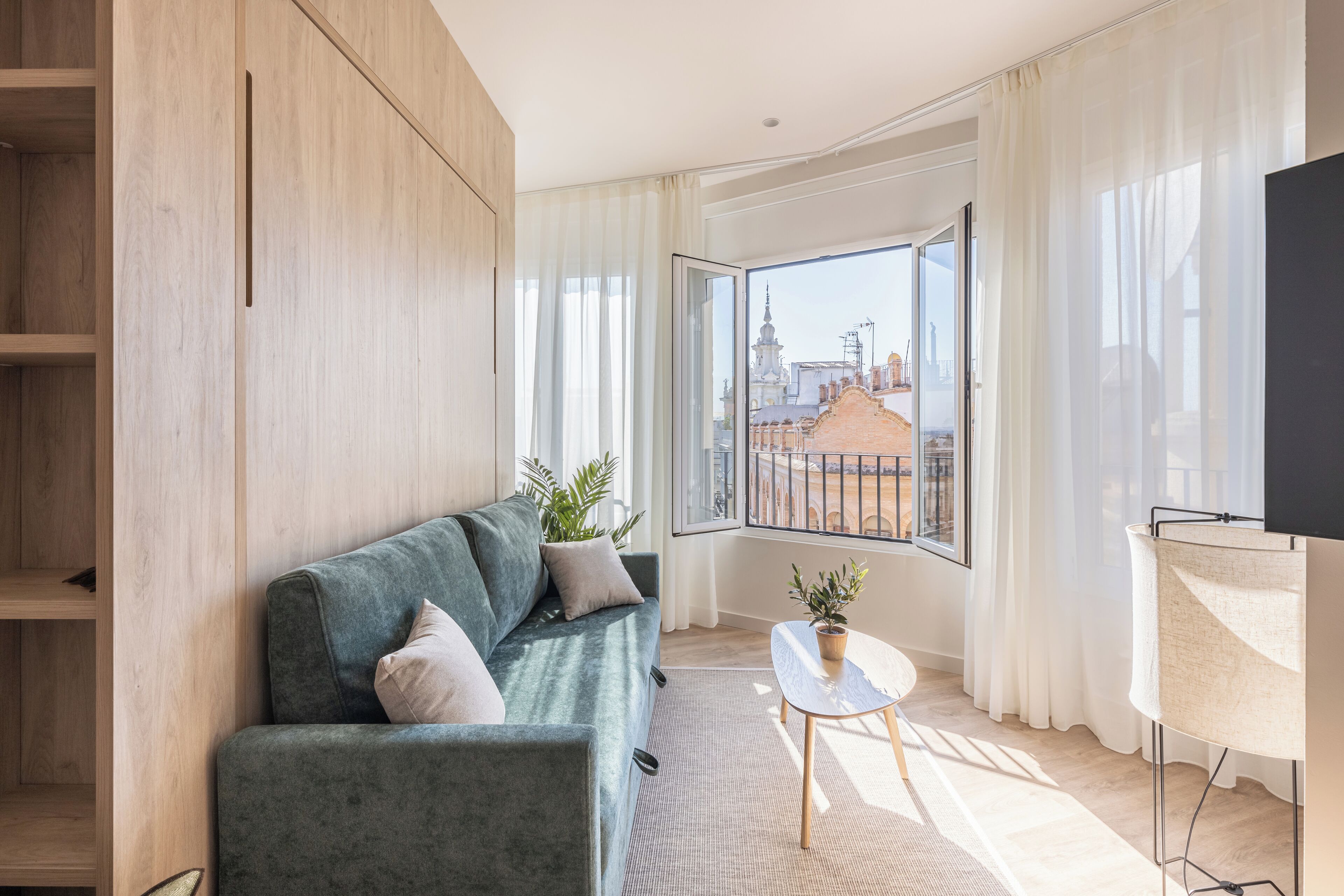 A modern living room with a grey sofa, a small table with a plant, and a large window offering a city view.
