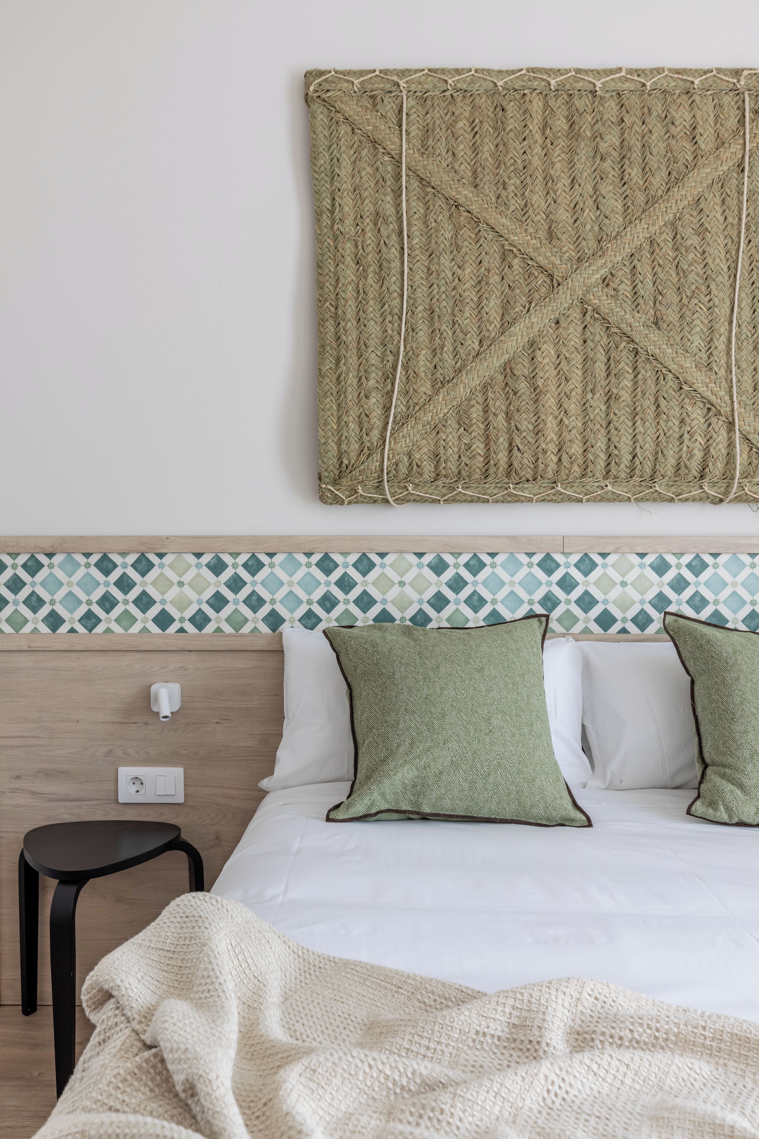 A neatly made bed with white and green pillows, a woven wall hanging above, and a small black stool beside it.