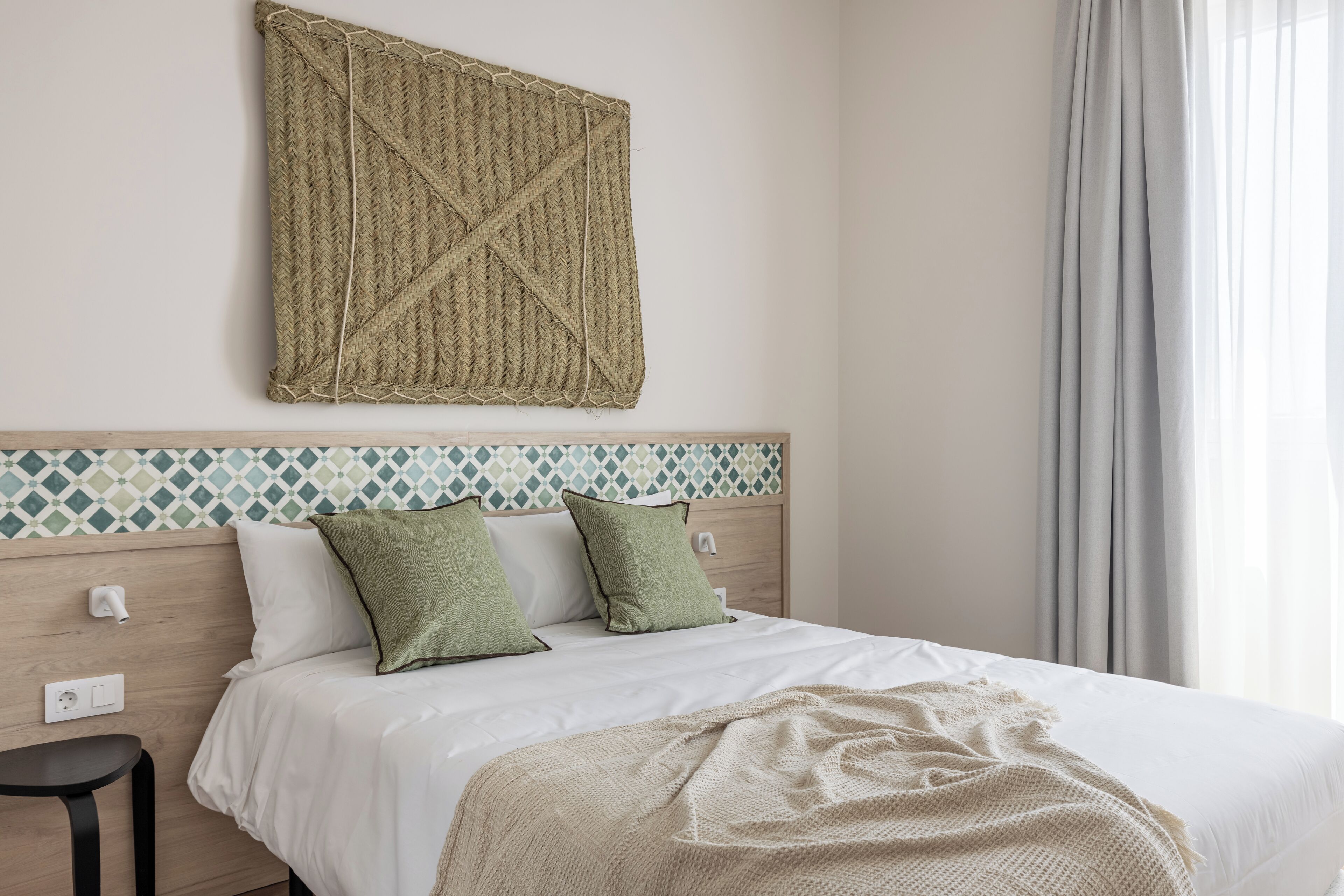 A neatly made bed with white bedding and two green pillows, a woven tapestry above the bed, and a patterned tile backsplash.