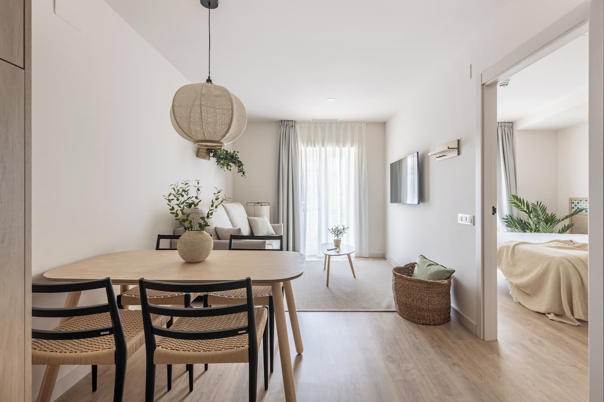 Une pièce moderne et bien éclairée, avec une table de salle à manger ronde, deux chaises, un canapé, un téléviseur fixé au mur et un lit recouvert d’une couverture beige.