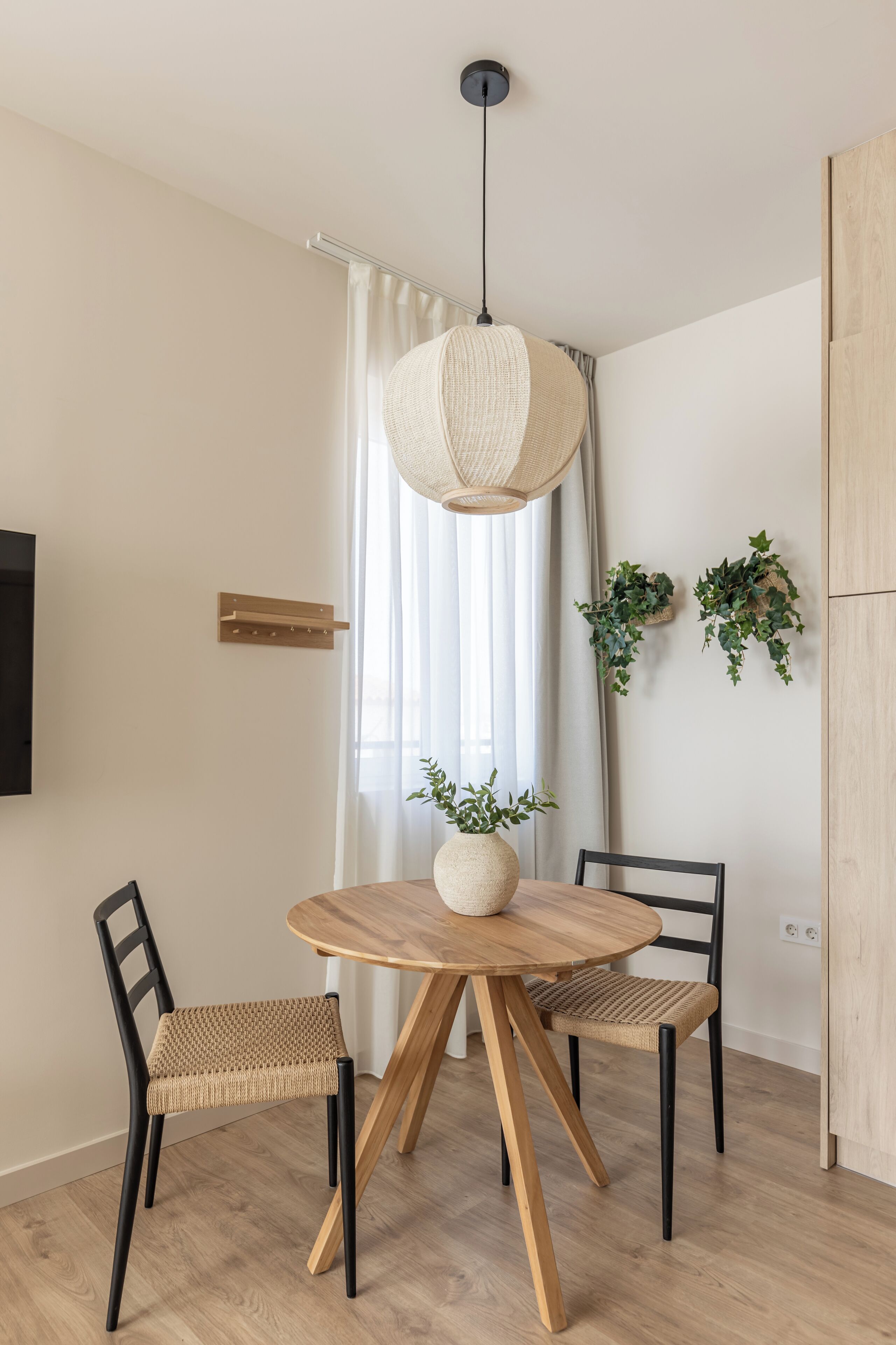Un coin repas comprenant une table ronde en bois, deux chaises noires, un luminaire suspendu en fibres tressées et une plante en pot.