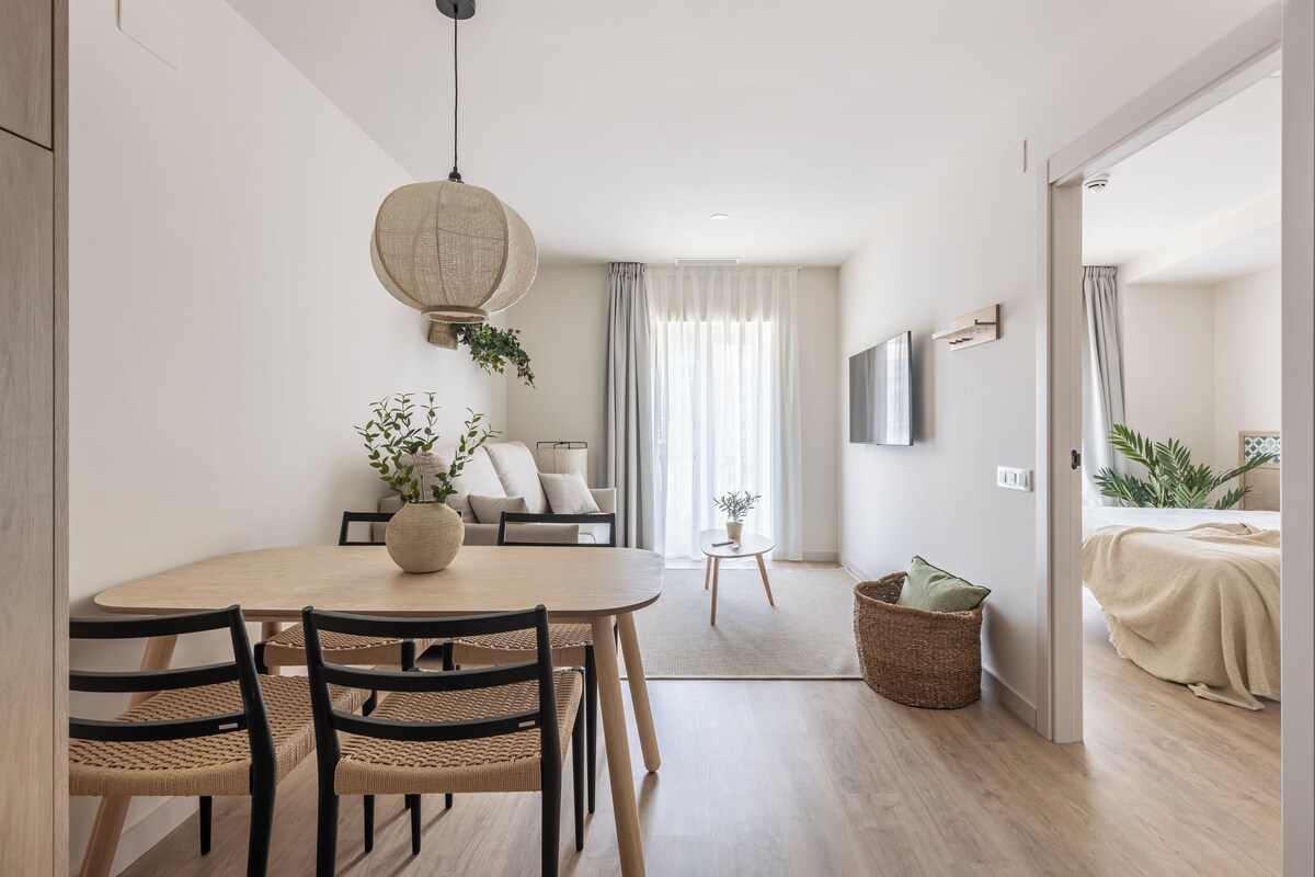 Una stanza moderna e luminosa con un tavolo rotondo, due sedie, un divano, una TV a parete e un letto con coperta beige.