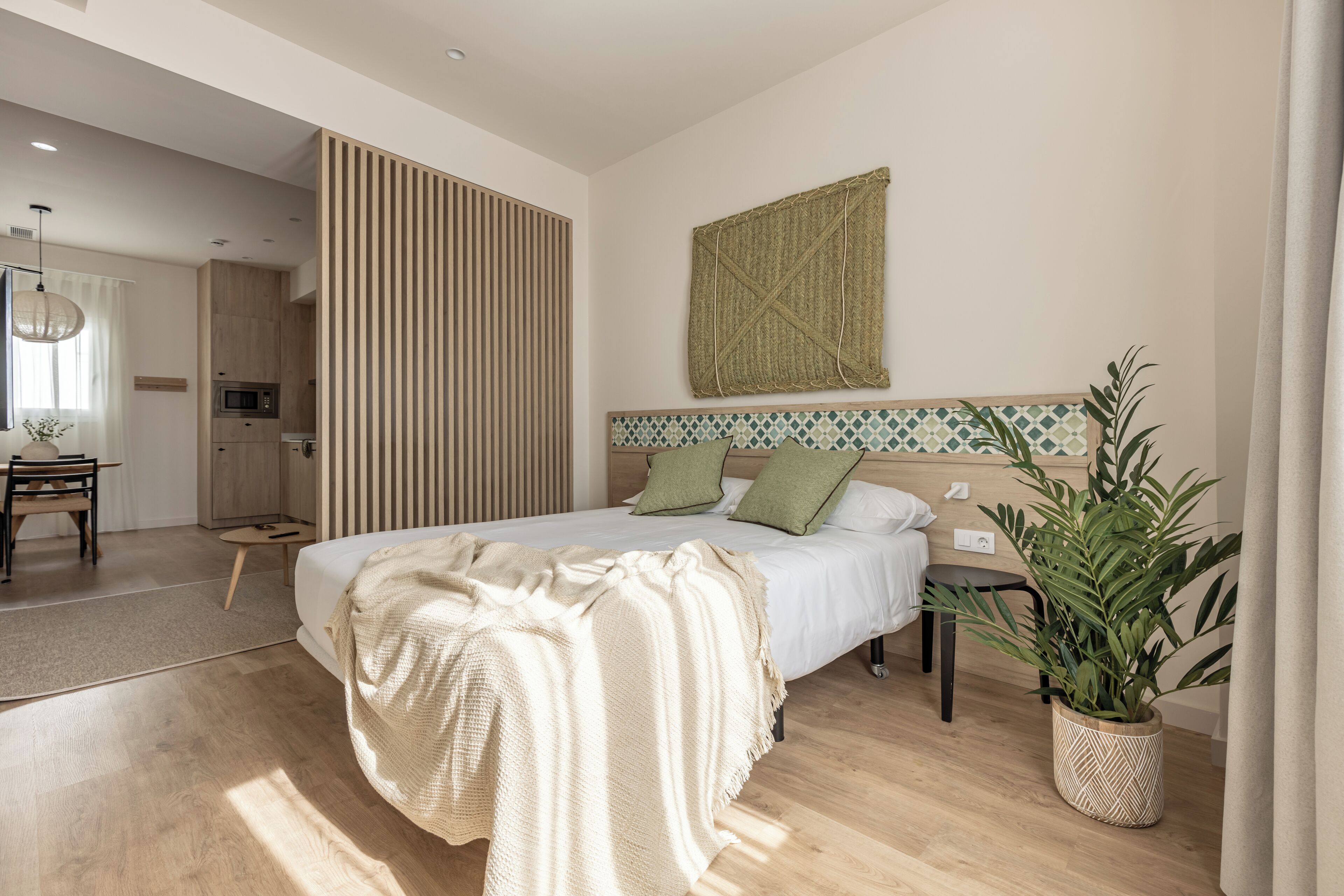 Quarto moderno com cama grande, piso de madeira, área de cozinha e um vaso com planta.
