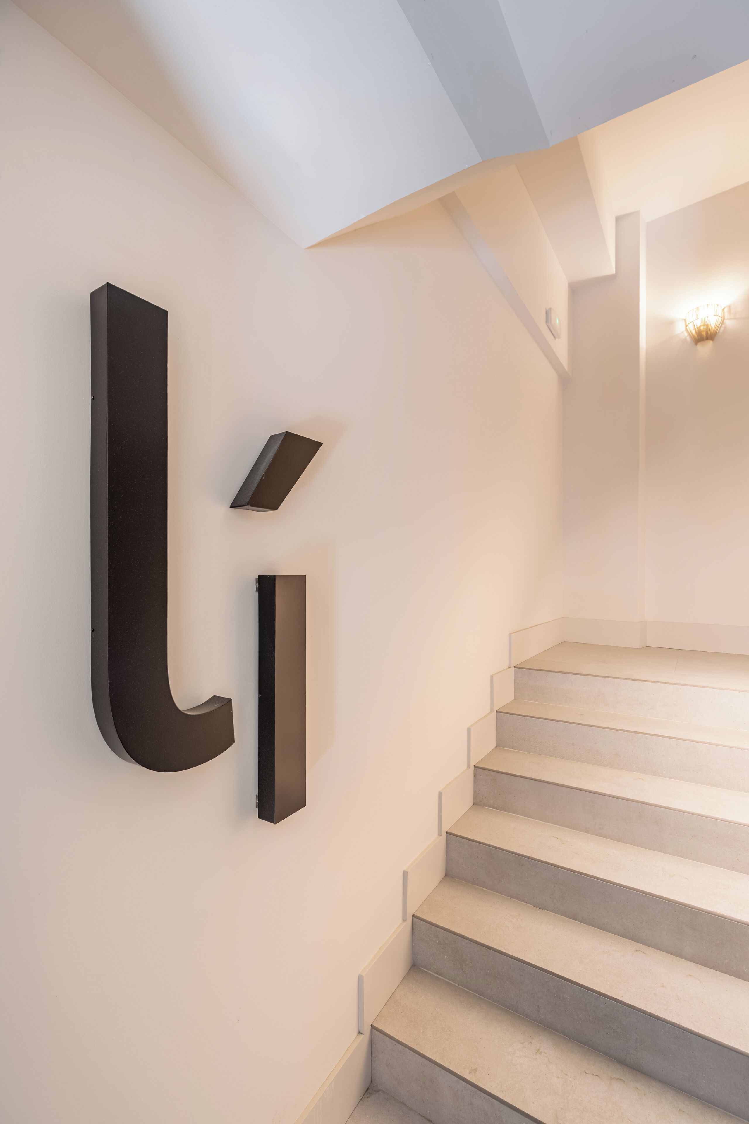 A modern staircase with a minimalist design, featuring a light-colored wall and a dark accent wall with a large, bold letter 'L'.