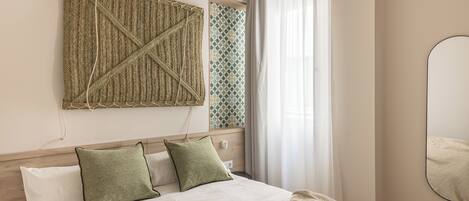 Una cama bien hecha con sábanas blancas y dos almohadas verdes, un tapiz de pared y un espejo.