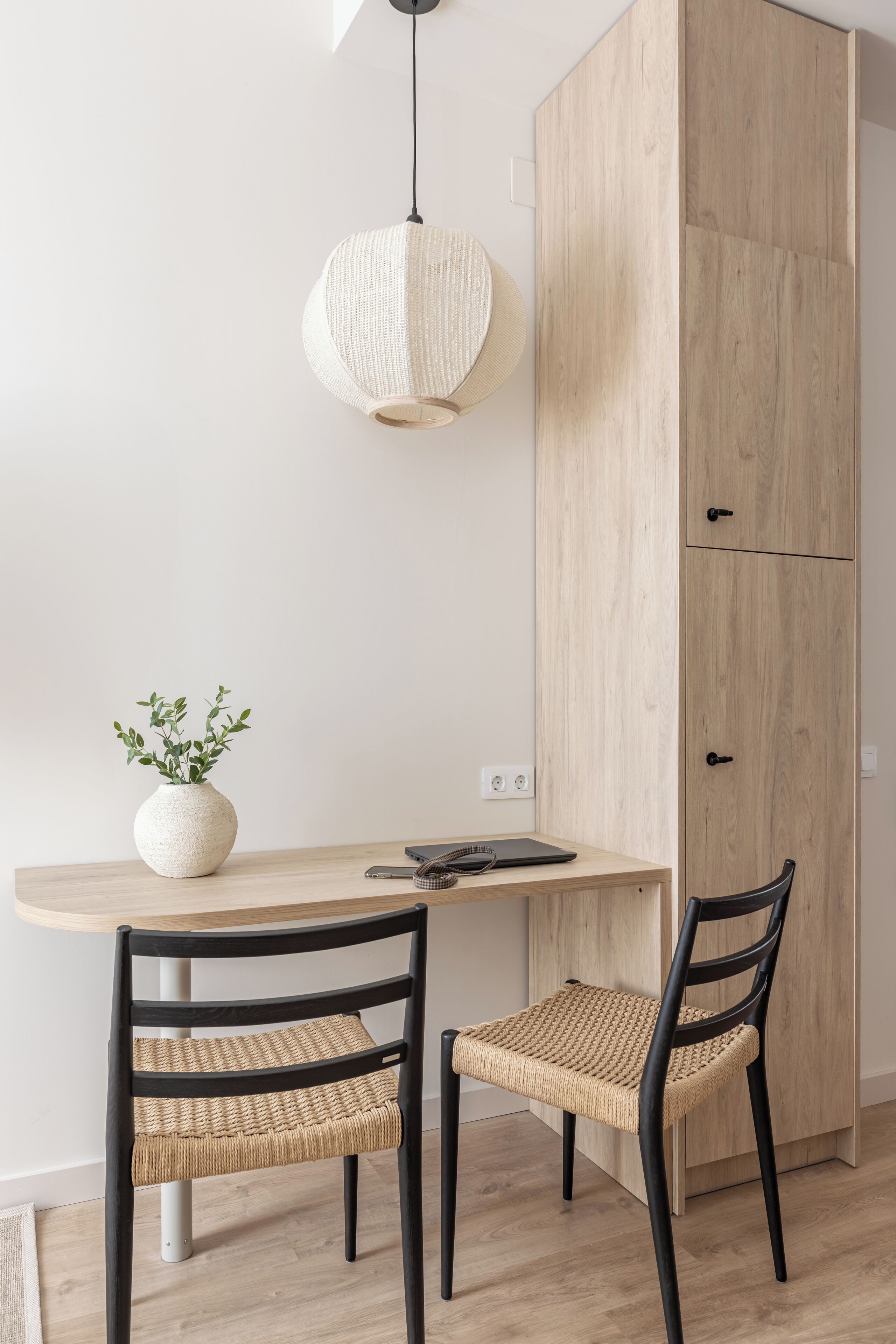 A dining area with a wooden table, two wicker chairs, a vase with green plants, and a hanging light fixture.