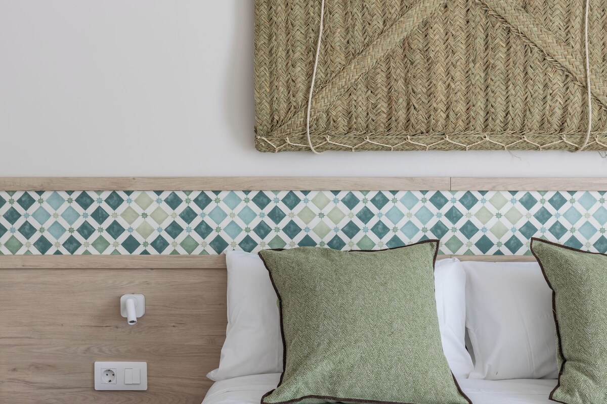 A neatly made bed with white and green pillows, a woven wall hanging above, and a small black stool beside it.