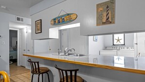 A kitchen with a central island, two bar stools, a stainless steel sink, and a large mirror. - Summer Winds 436B- 2BR Condo w/ Ocean Views & Pools! (Indian Beach)