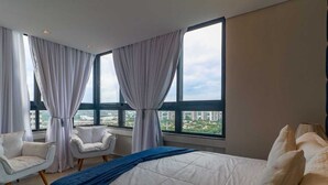 Une chambre d’hôtel avec un grand lit, deux fauteuils blancs, une petite table et une vue sur la ville grâce à de grandes fenêtres.