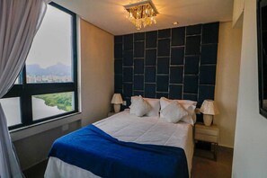A hotel room with a large bed, two bedside lamps, a view of a cityscape through a window, and a decorative chandelier.