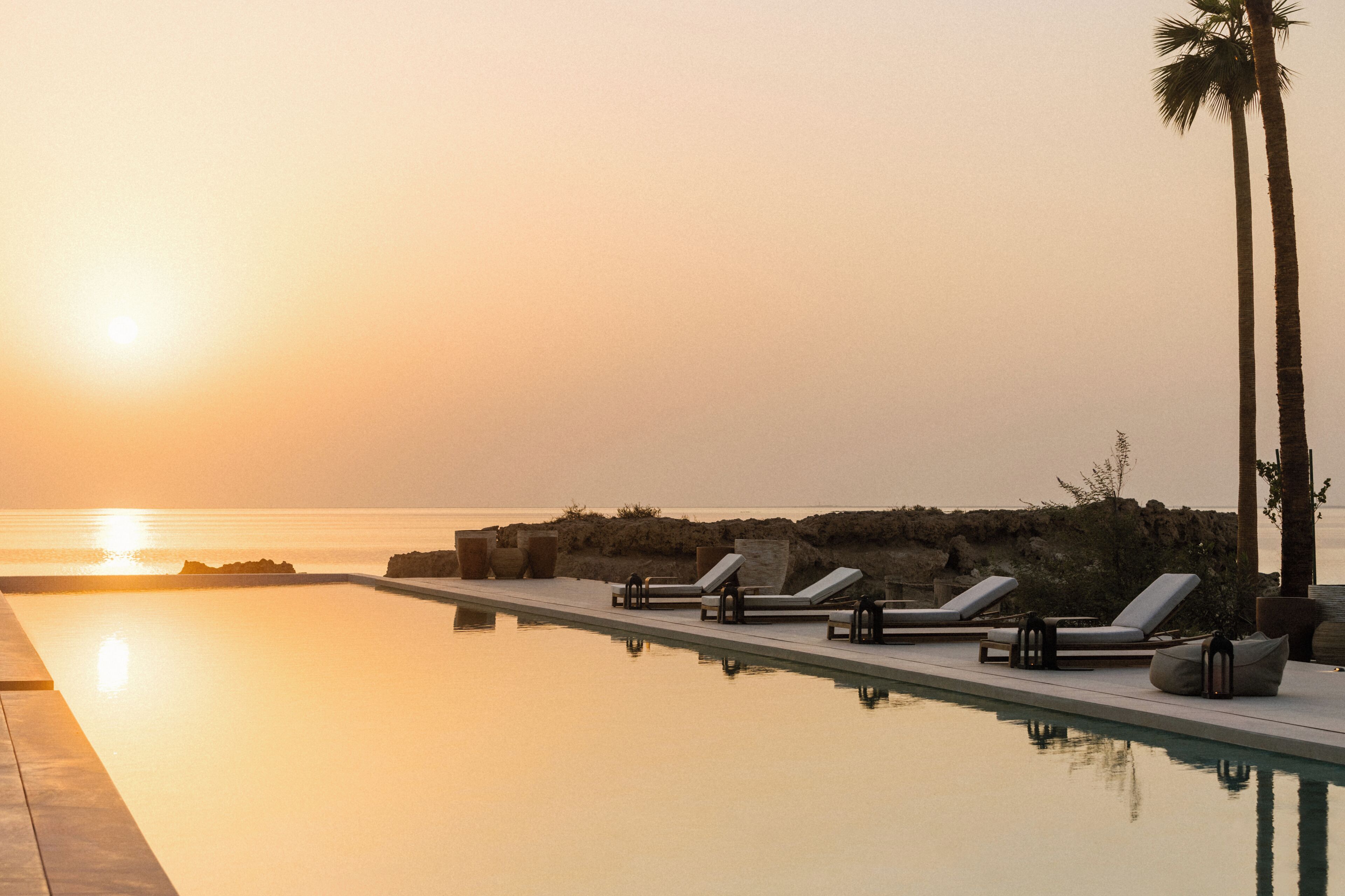 A poolside area with lounge chairs and a palm tree, overlooking the ocean at sunset.