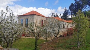A stone building with a red-tiled roof, surrounded by blooming trees and a stone wall in the foreground. - Solar dos Condes da Azenha (Sernancelhe)